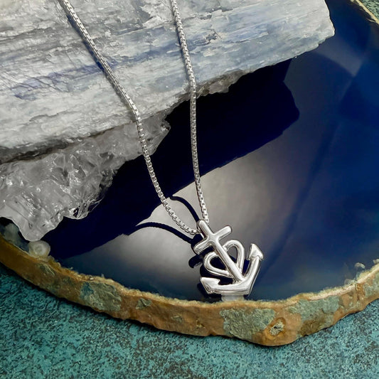 A silver necklace with a pendant featuring an anchor symbol is displayed on a blue and green textured surface.