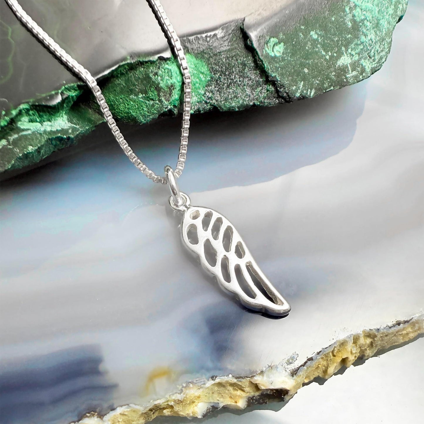A silver-colored pendant with a wing design is displayed on a chain, resting on a green and gray rock formation.