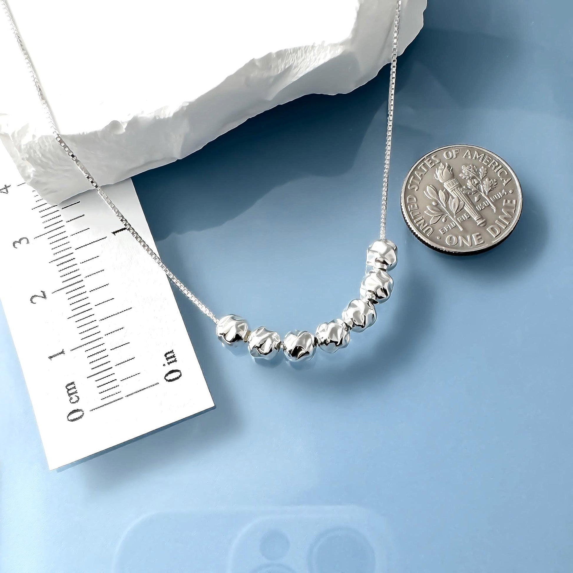 a necklace with a row of silver beads, a ruler for scale, and a United States quarter coin for size comparison.