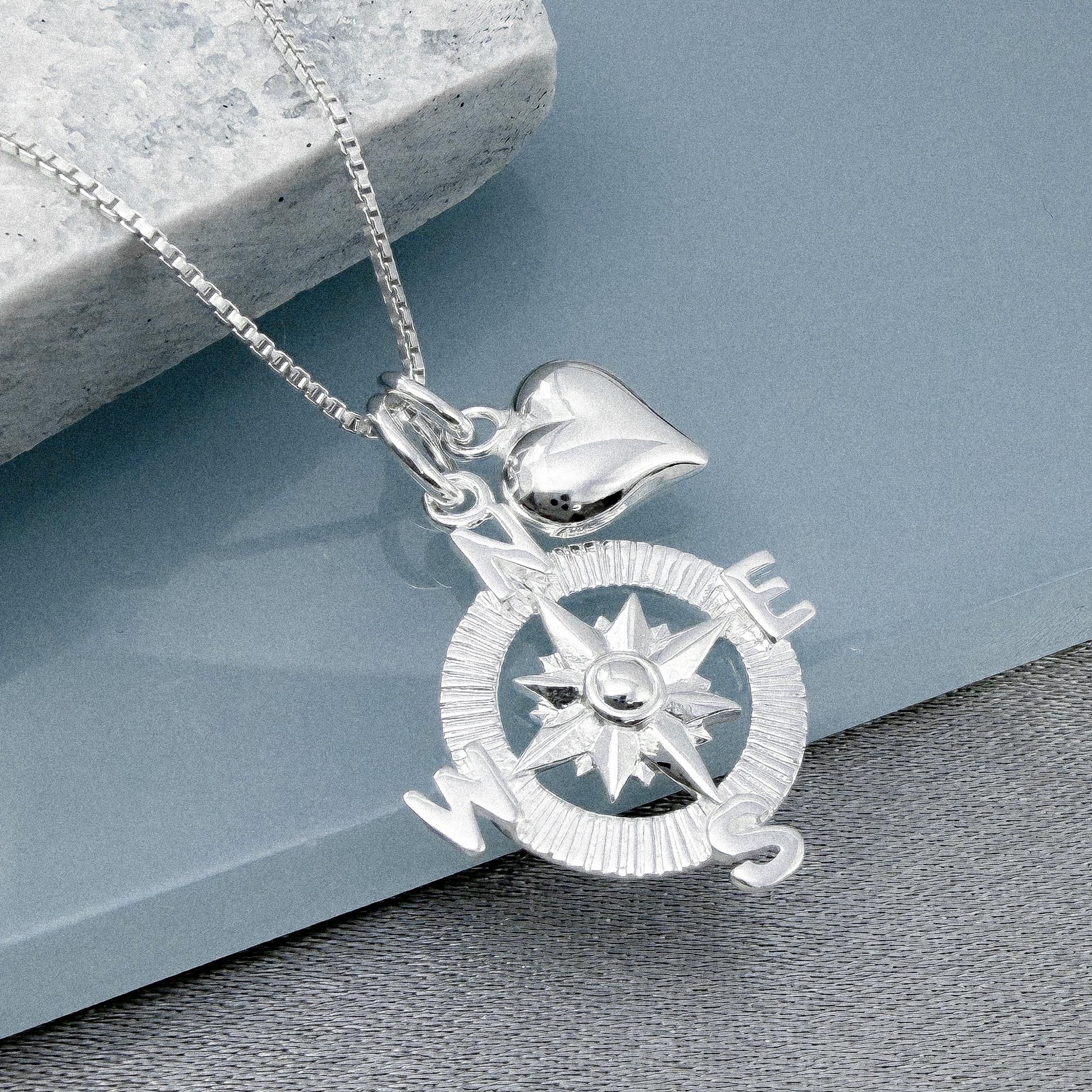 A silver compass pendant with a heart charm attached, resting on a gray surface.