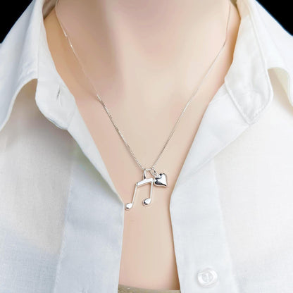 A silver necklace with a heart-shaped charm and musical notes pendant is displayed on a person wearing a white collared shirt.