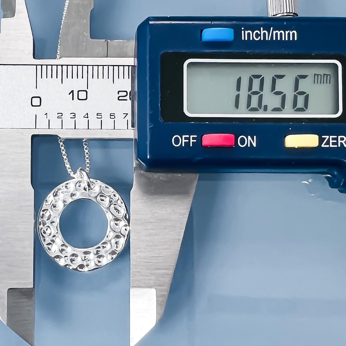 A silver necklace with a circular pendant is being measured against a blue scale, which displays the weight of the necklace as 18.56 millimeters.