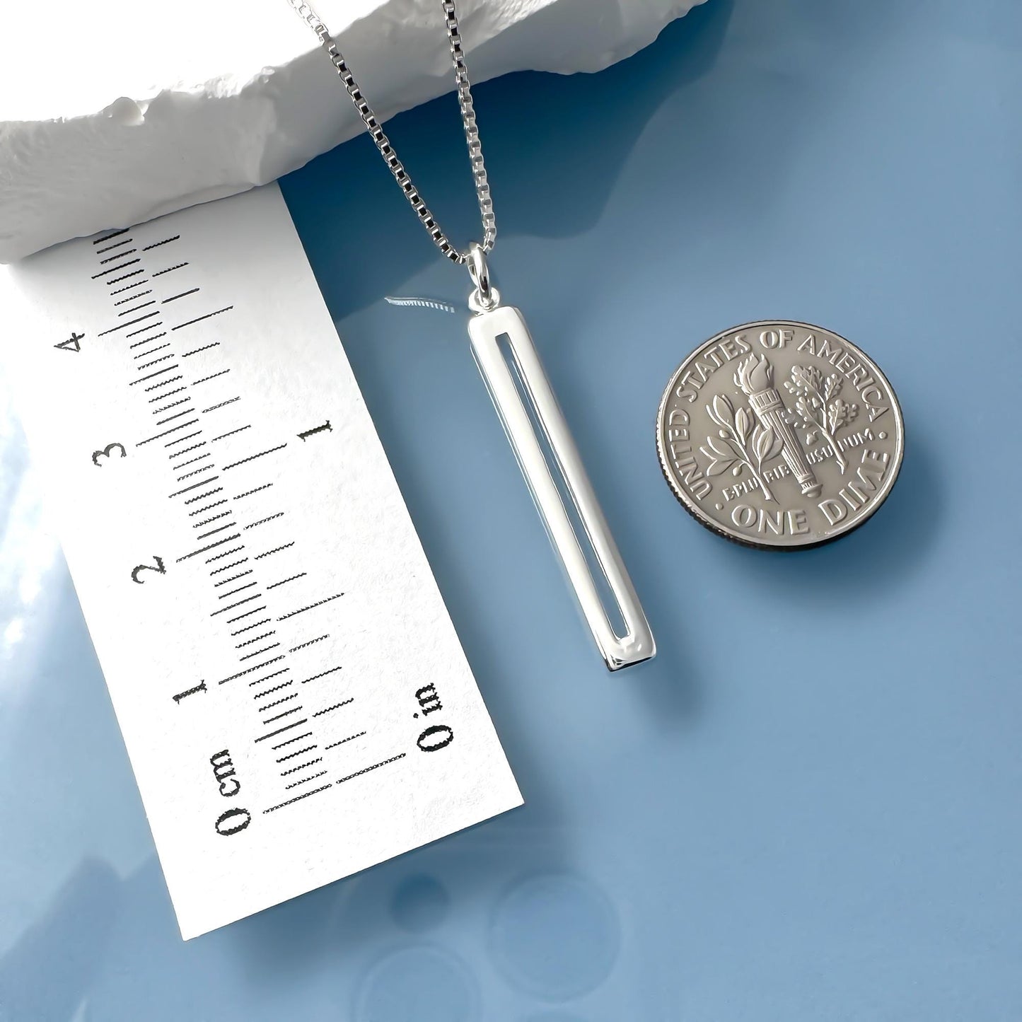 a silver necklace with a pendant, a ruler, and a United States one dollar coin on a blue surface.