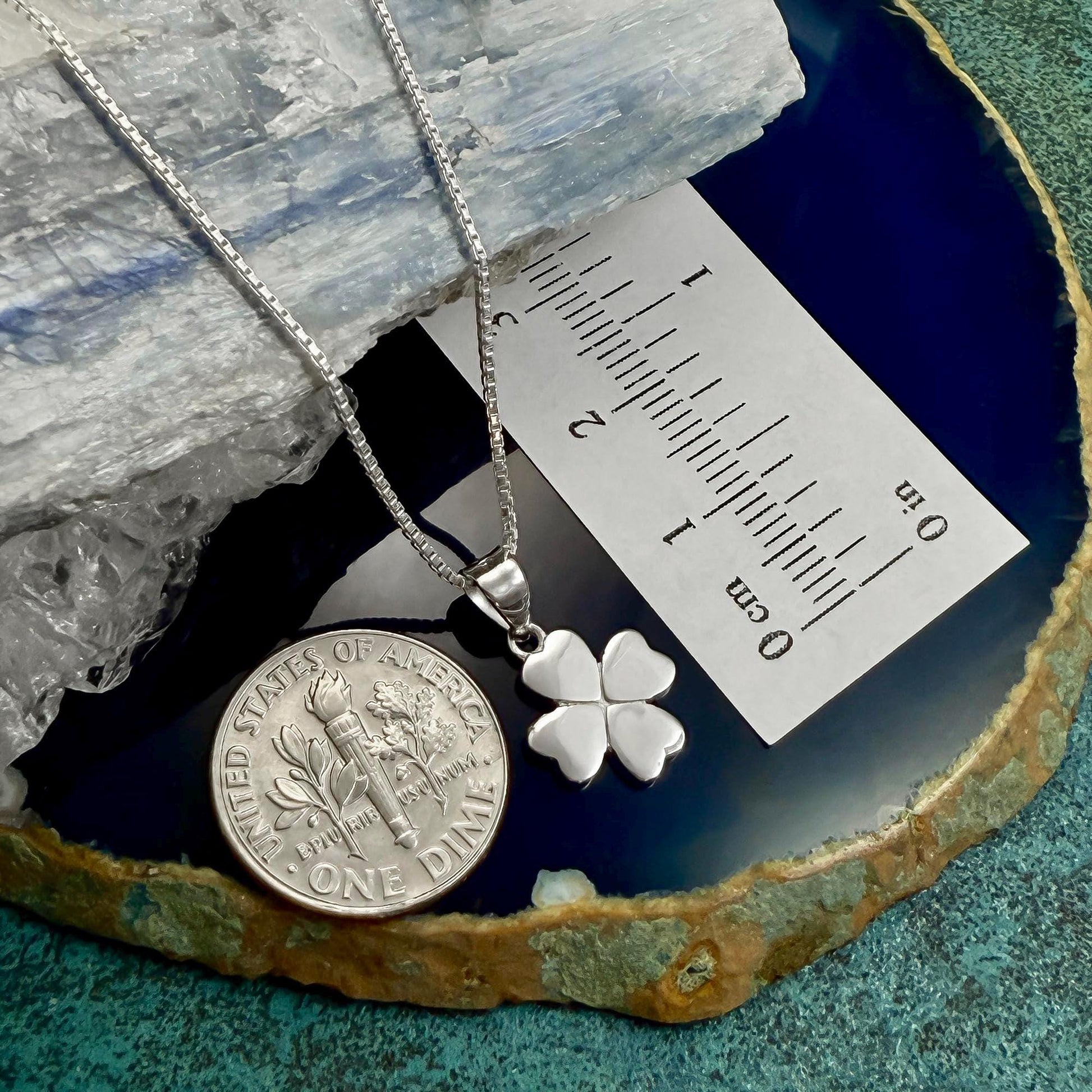 a silver necklace with a four-leaf clover pendant, placed on a blue and green surface. Next to the necklace is a silver coin, and there is a ruler placed on top of the coin.