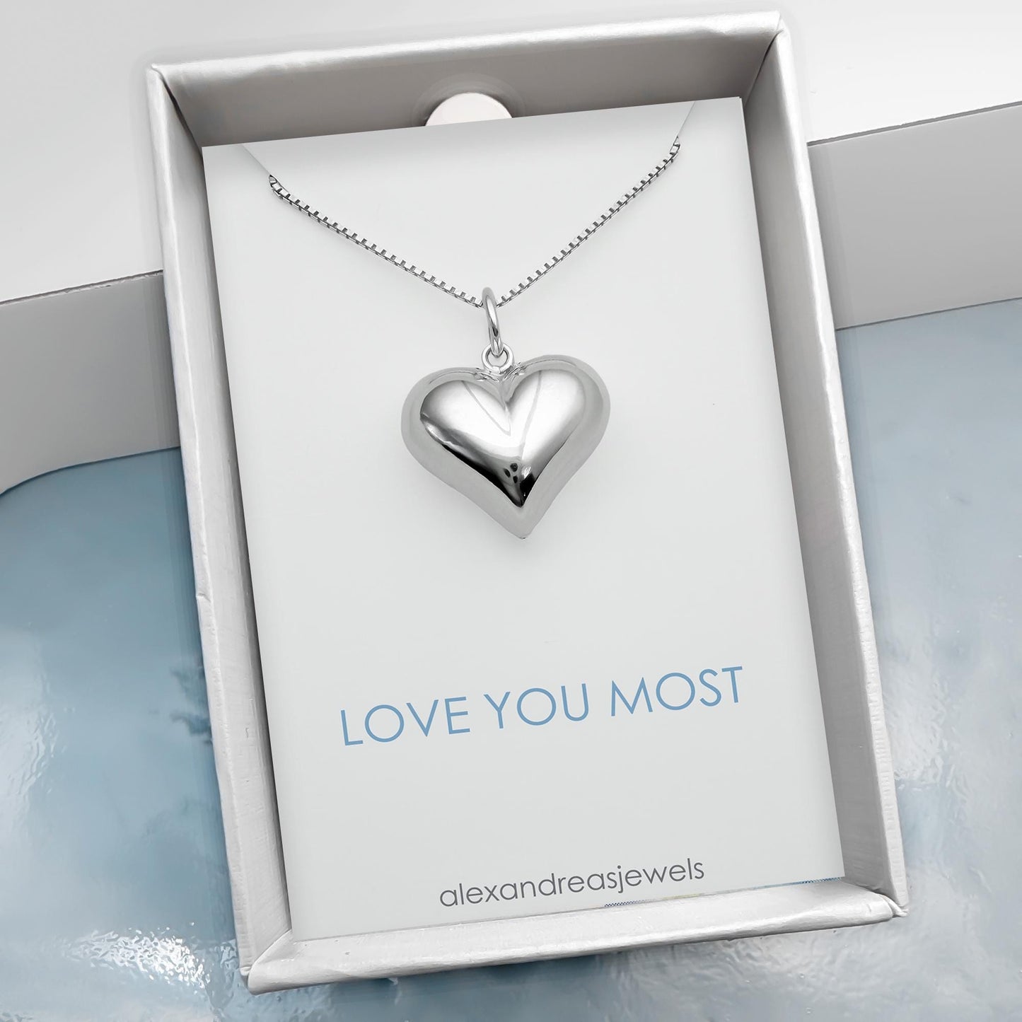 A silver heart-shaped pendant necklace is displayed in a white box with the text "LOVE YOU MOST" and "alexandreajels" printed on it.