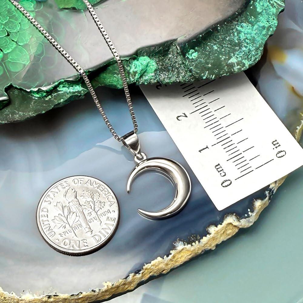 A silver necklace with a crescent moon pendant is displayed next to a United States one dollar coin, which is lying on a blue and green rock formation.